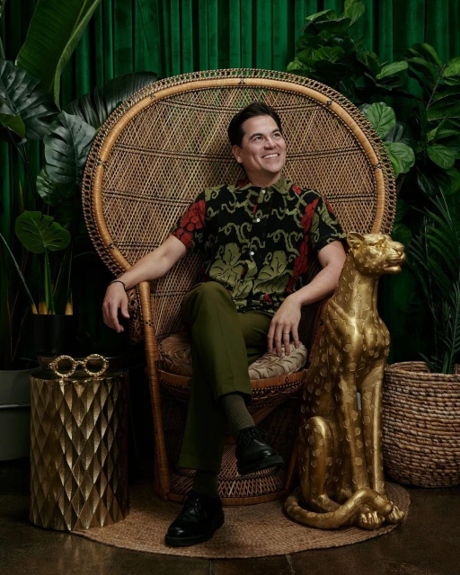 Stylish man sits in a peacock chair surrounded by tropical foliage, a golden panther statue, and a geometric side table with sunglasses.